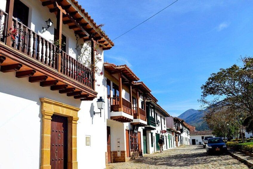 A cobblestone street lined with traditional buildings, a car parked on the right, and mountains in the background. 3