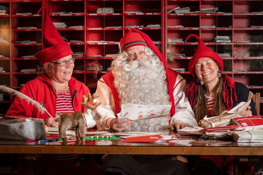 Three people in Santa hats, a table with wrapping paper, and a background of red shelves with mail. 1