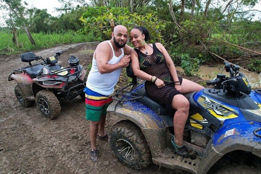 Zwei Personen fahren mit einem blauen Quad durch eine schlammige Gegend, im Hintergrund ist ein weiteres Quad zu sehen. 2