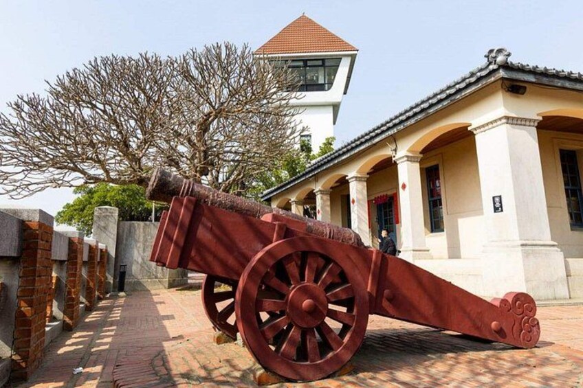 A large cannon with a red barrel and wheels, positioned in front of a building with a tiled roof and columns. 5