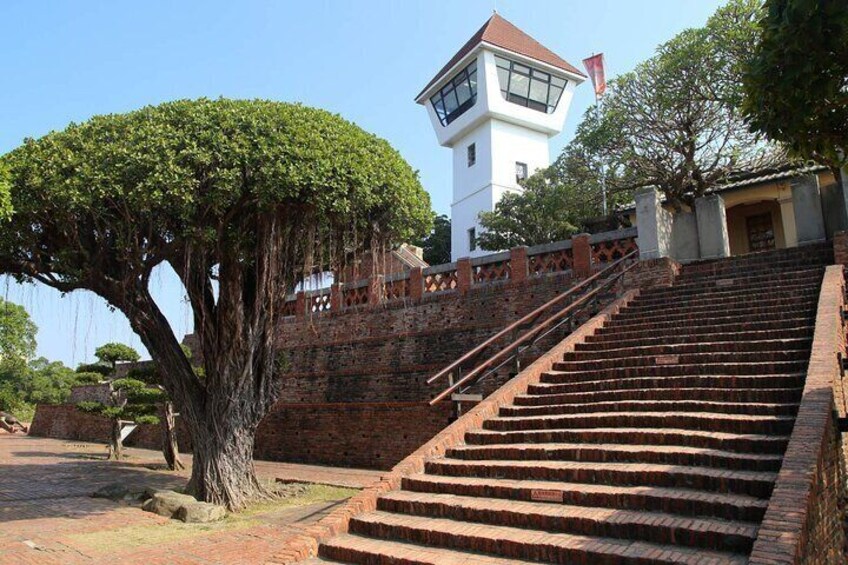 A staircase leading up to a white tower with a red flag, surrounded by trees and brick walls. 1