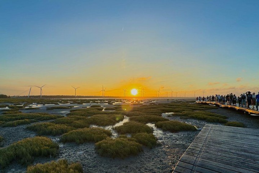 Un coucher de soleil sur une zone côtière, avec des éoliennes et une promenade en bois où se promènent des gens. 1