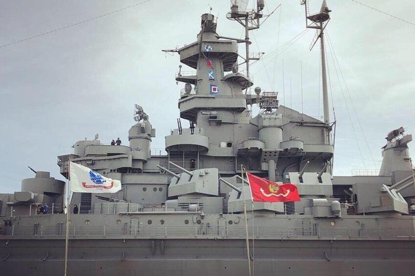 A large naval ship with multiple flags, including a red flag with a yellow emblem, and a white flag with a blue emblem. 1