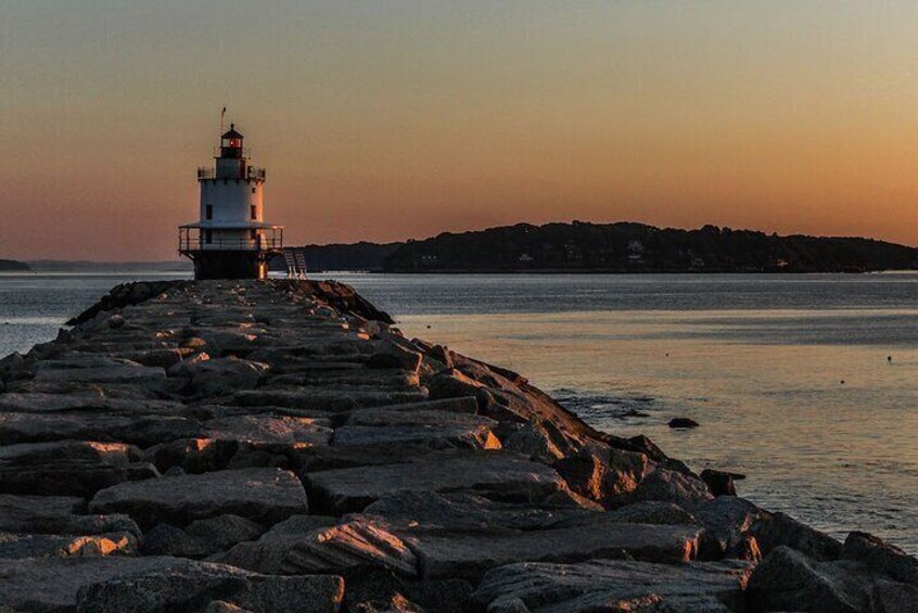 A lighthouse on a rocky pier extending into calm waters, with a sunset in the background. 2