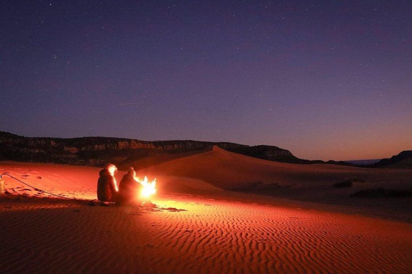 A desert landscape at night with a campfire and two people sitting near it. 4