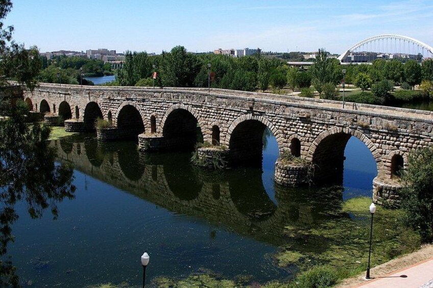 A stone bridge over a calm river, surrounded by greenery and a distant stadium. 4