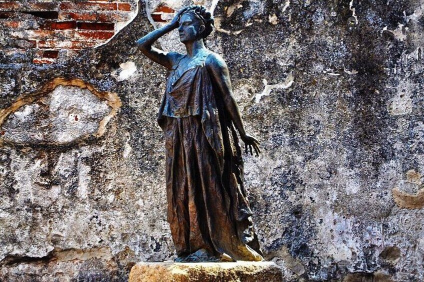 A weathered statue of a woman with a draped cloak, set against a brick wall with visible wear and tear. 2