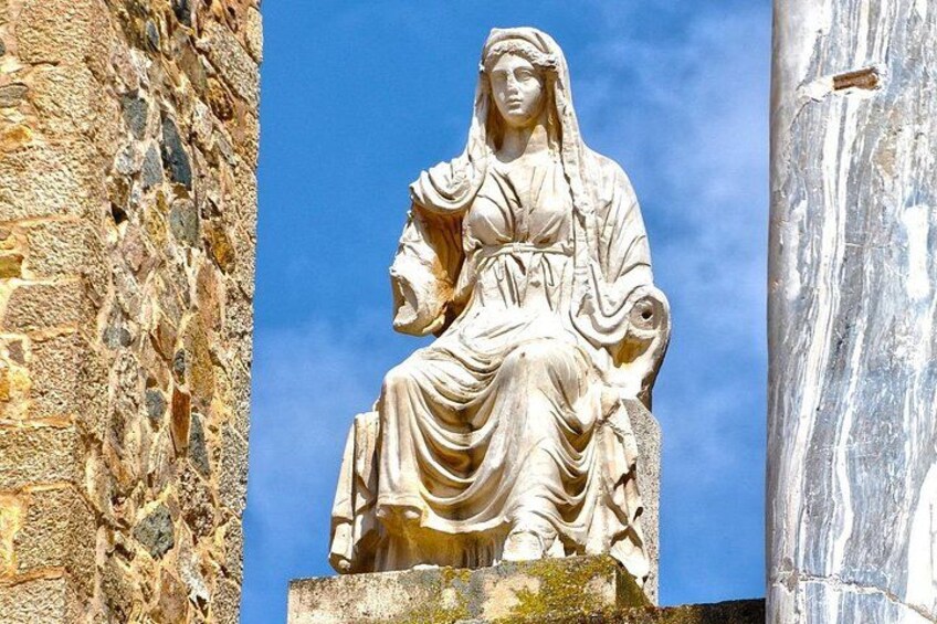 A stone statue of a seated woman with a horse, set against a clear blue sky, with a stone wall and a marble column in the background. 3