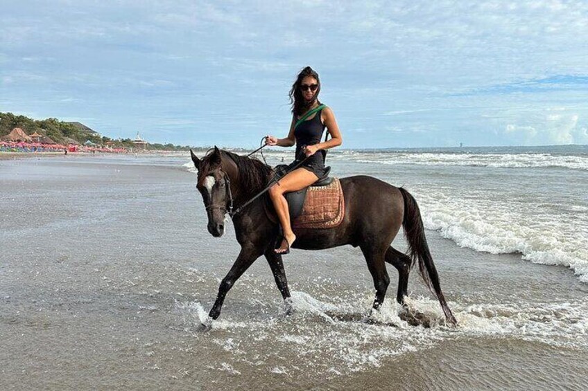 Una persona montando a caballo en una playa con olas, cielo despejado y edificios al fondo. 4