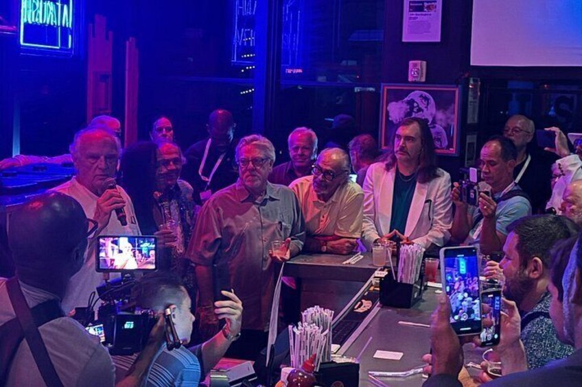 A group of people gathered around a table with a microphone, some holding drinks, and a person speaking into the microphone. 4