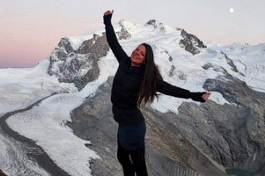 Eine Person steht in einer Berglandschaft mit schneebedeckten Gipfeln und streckt die Arme in die Höhe. 3
