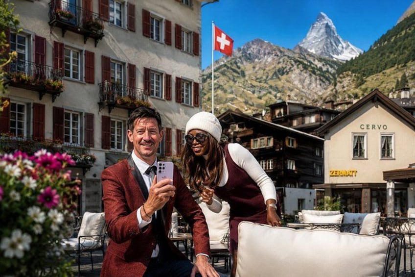 Ein Paar macht ein Foto mit einem Smartphone, im Hintergrund weht eine Schweizer Flagge und ein Gebäude mit der Aufschrift 'CERVO' ist zu sehen. 2