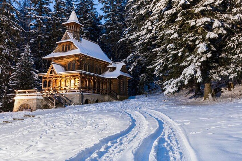 A snow-covered wooden cabin with a steep roof, surrounded by snow-laden trees and a winding path. 5
