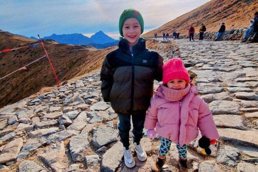 A rocky path with a person and a child standing on it, mountains in the background, and a few people in the distance. 4