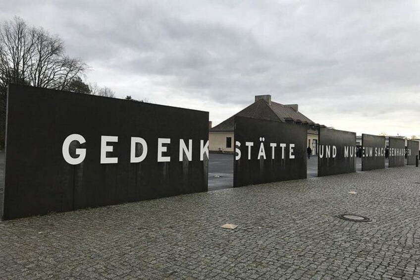 A paved area with a large black wall displaying the text 'GEDENKSTÄTTE' and a building in the background. 3
