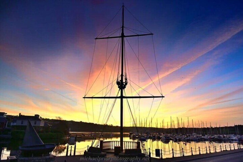 Un voilier à mât élevé et aux cordages complexes, amarré dans une marina, avec d'autres bateaux en arrière-plan, au moment du coucher du soleil. 5