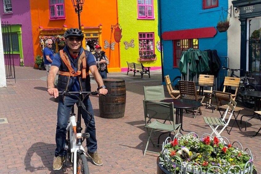 A person on a bicycle in a colorful street with outdoor seating and a flower arrangement. 1