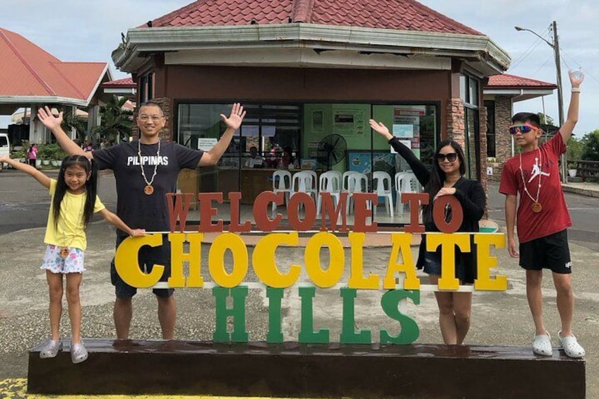 Een groep mensen staat voor een bord waarop 'WELCOME TO CHOCOLATE HILLS' staat. 2