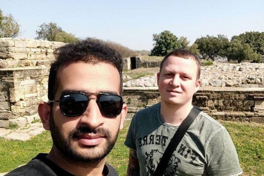 Dos personas frente a ruinas antiguas con estructuras de piedra y vegetación al fondo. 3