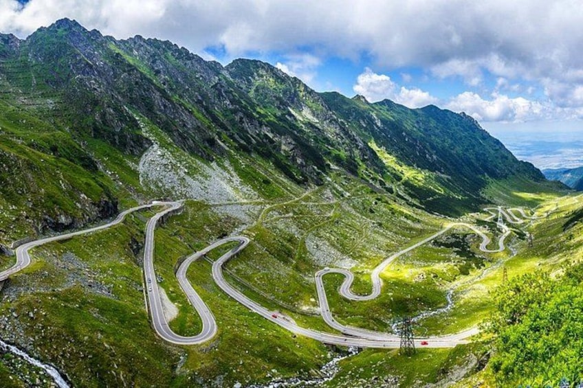 A winding mountain road surrounded by lush green hills and rocky mountains under a partly cloudy sky. 1