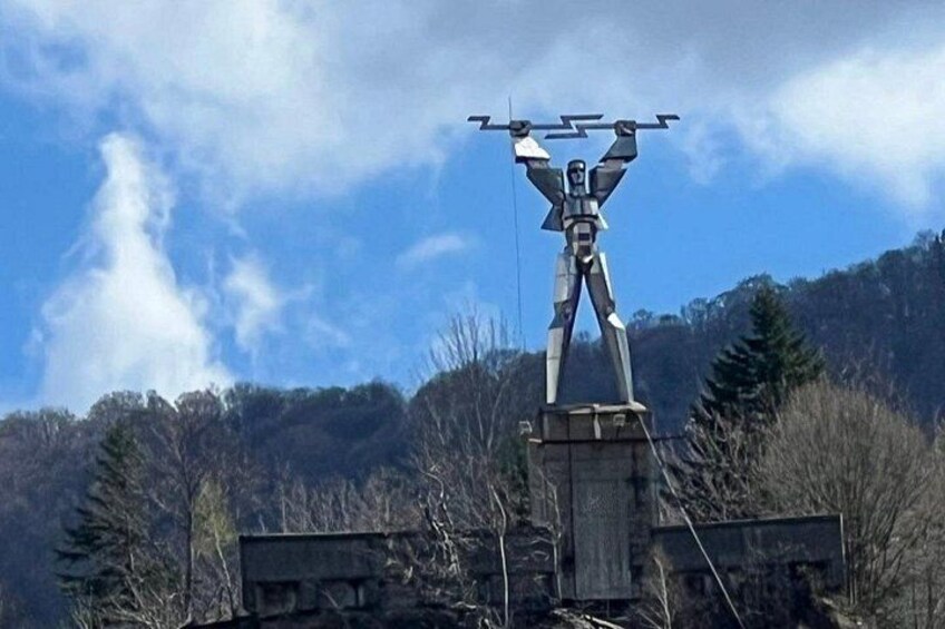 A sculpture of a humanoid figure with a drone on its head, set against a backdrop of trees and a mountain. 4