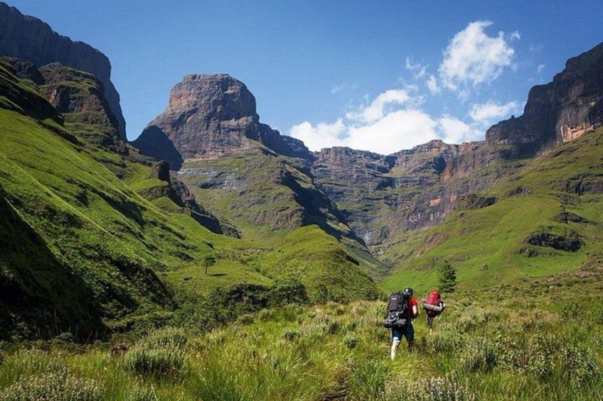 A hiker with a backpack and red backpack walking on a grassy hillside with mountains in the background. 2