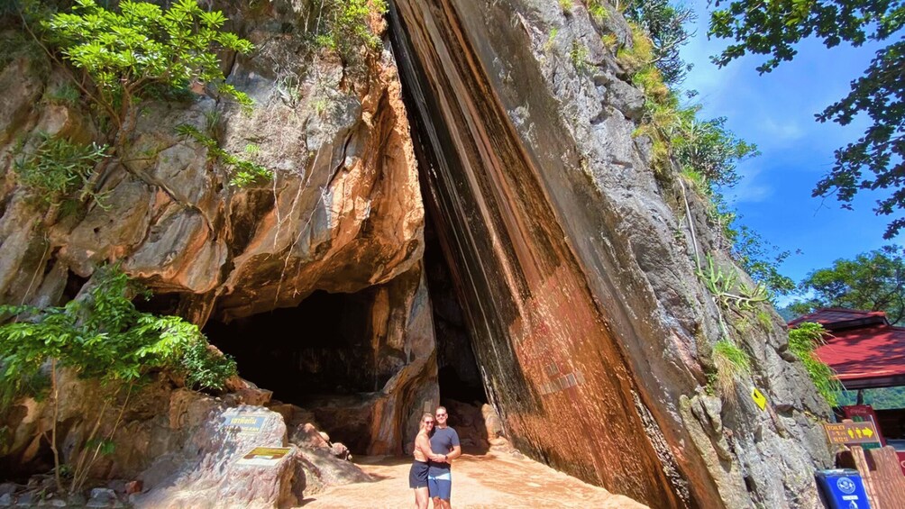 A cave entrance with a person standing in front, surrounded by rocky cliffs and greenery. 5