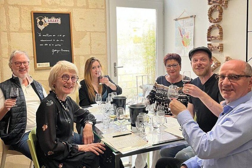 A group of people sitting around a table with wine glasses, a framed quote on the wall, and a window in the background. 2