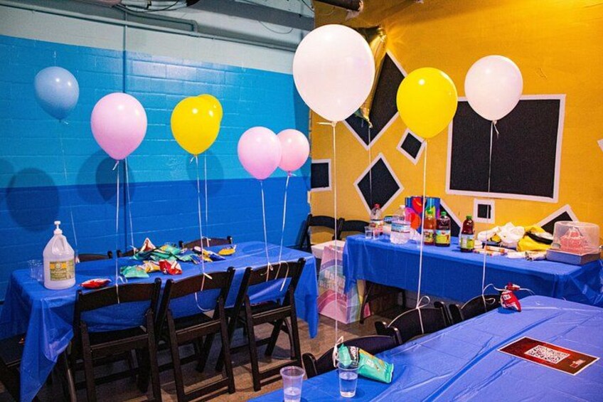 Una habitación con mesas cubiertas con manteles azules, globos de varios colores y artículos para fiestas. 2