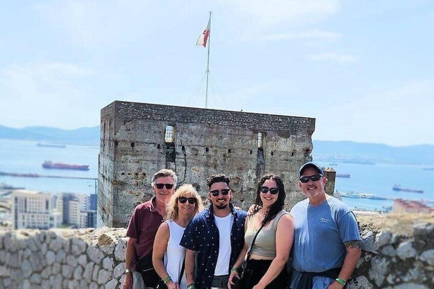 Un grupo de personas frente a una estructura histórica de piedra con una bandera, con vista a una masa de agua y barcos a lo lejos. 2