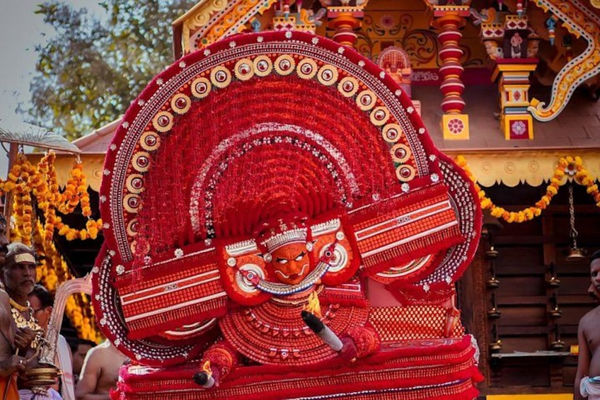 Una estatua roja grande y ornamentada, con diseños intrincados y un motivo circular, ubicada frente a un templo de arquitectura detallada. 2