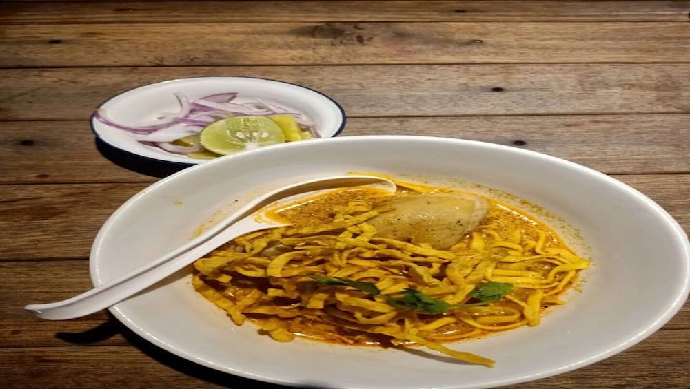 A bowl of yellow noodles with a spoon, garnished with cilantro, and a small bowl of sliced onions and lime. 2
