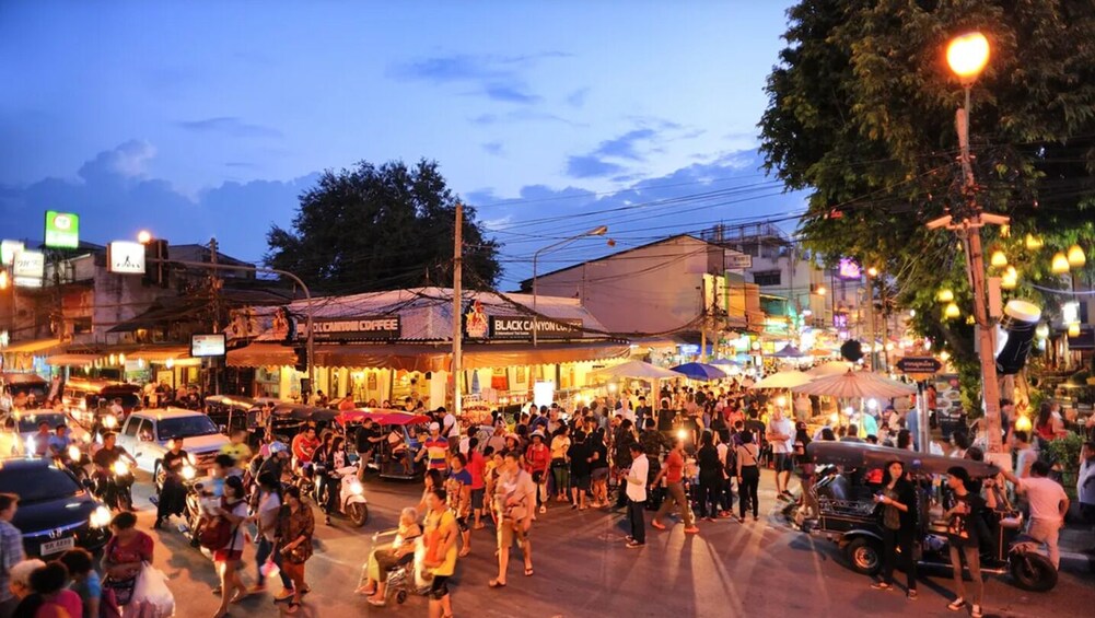 A bustling street market at dusk with people walking and vehicles parked along the roadside. 1