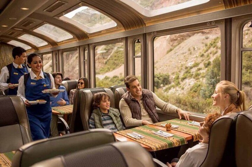 A train dining car with passengers seated at tables, a waiter serving food, and a mountainous landscape visible through the windows. 1