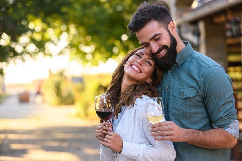 A couple holding wine glasses, standing outdoors with trees and a building in the background. 2