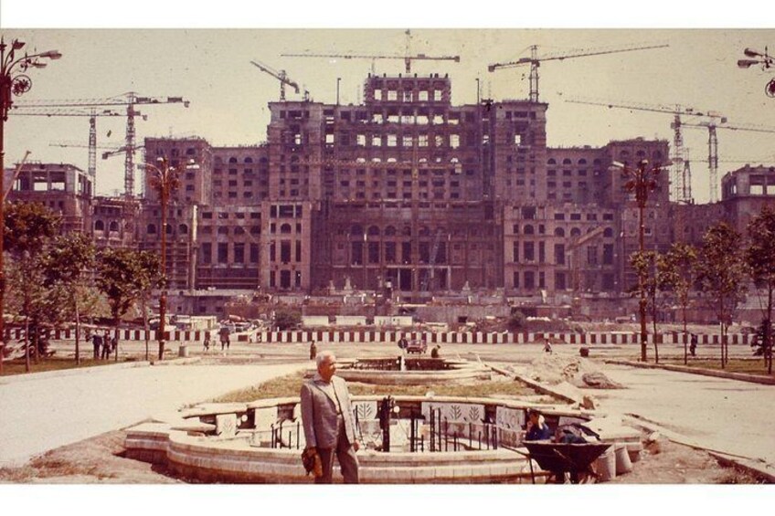 Un edificio grande en construcción con varias grúas, una fuente y un hombre de pie al frente. 1