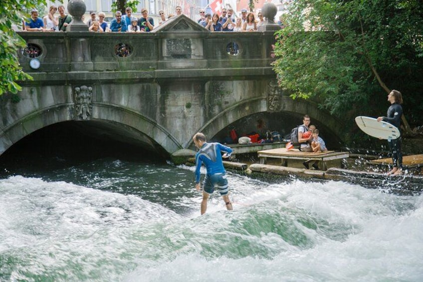 Une personne pratiquant le surf sur une rivière, un pont de pierre et des spectateurs qui observent depuis une plateforme à proximité. 1