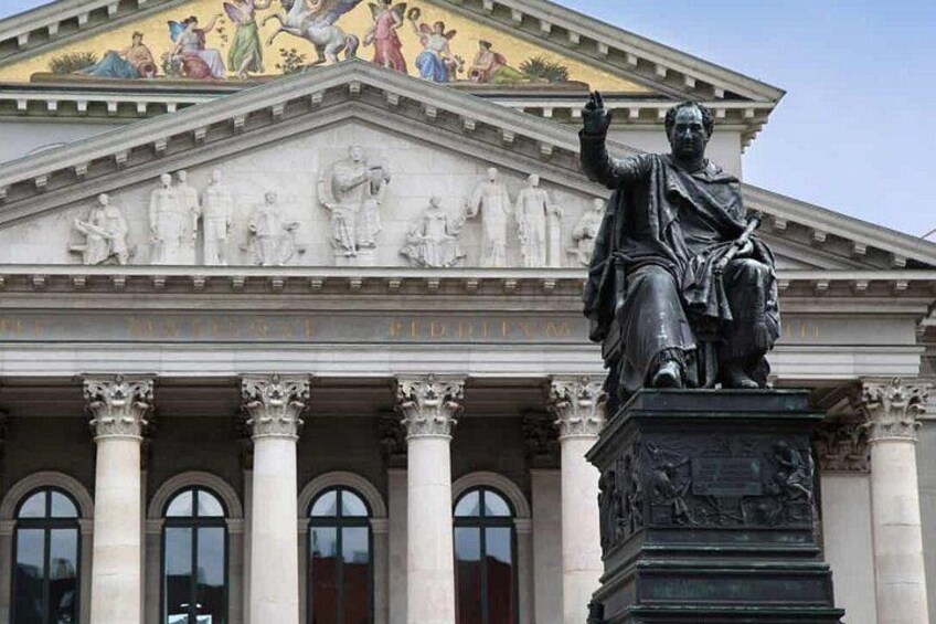 Eine klassische Gebäudefassade mit einer Statue eines sitzenden Mannes, Säulen und einem detaillierten Fries. 3