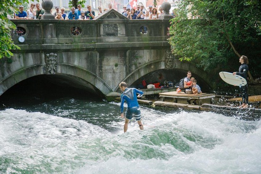 Eine Person surft auf einem Fluss, eine Steinbrücke und Zuschauer beobachten vom nahegelegenen Steg. 1