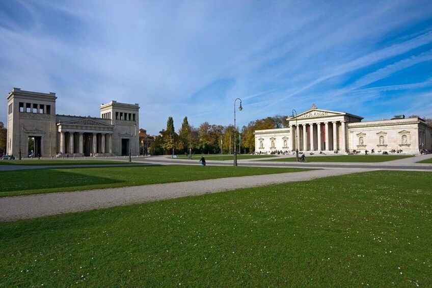 Ein großes, klassizistisches Gebäude mit Säulen und einem zentralen Giebel, umgeben von einer gepflegten Rasenfläche und blauem Himmel. 2