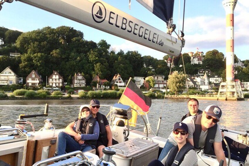 Un grupo de personas en un velero con una bandera alemana, un faro y casas a lo largo de la ribera del río. 1