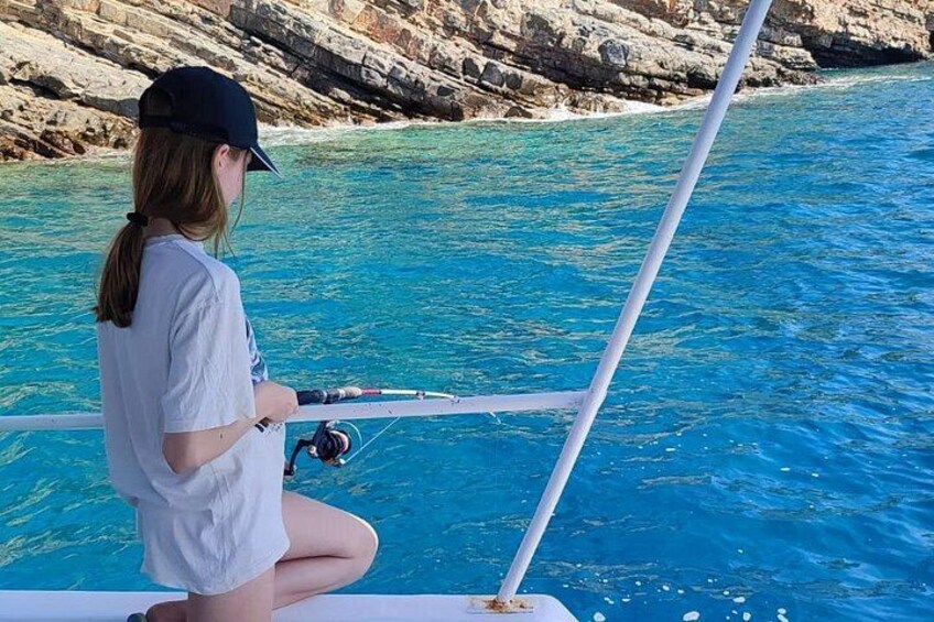 A person fishing on a boat with clear blue water and rocky coastline. 2