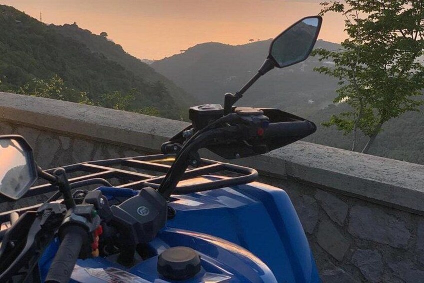 En blå motorcykel parkeret på en klippeafsats med udsigt over et bjergrigt landskab i solnedgang. 4