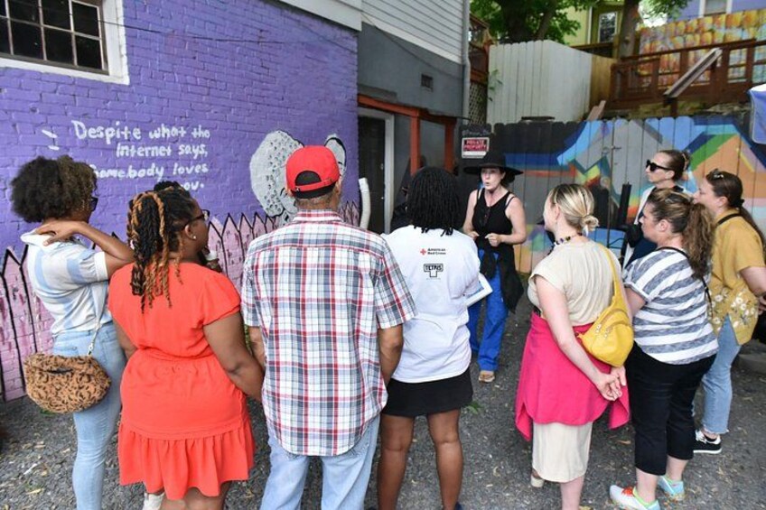 A group of people standing in front of a purple building with a mural, a man in a plaid shirt, and a woman in a red dress. 2