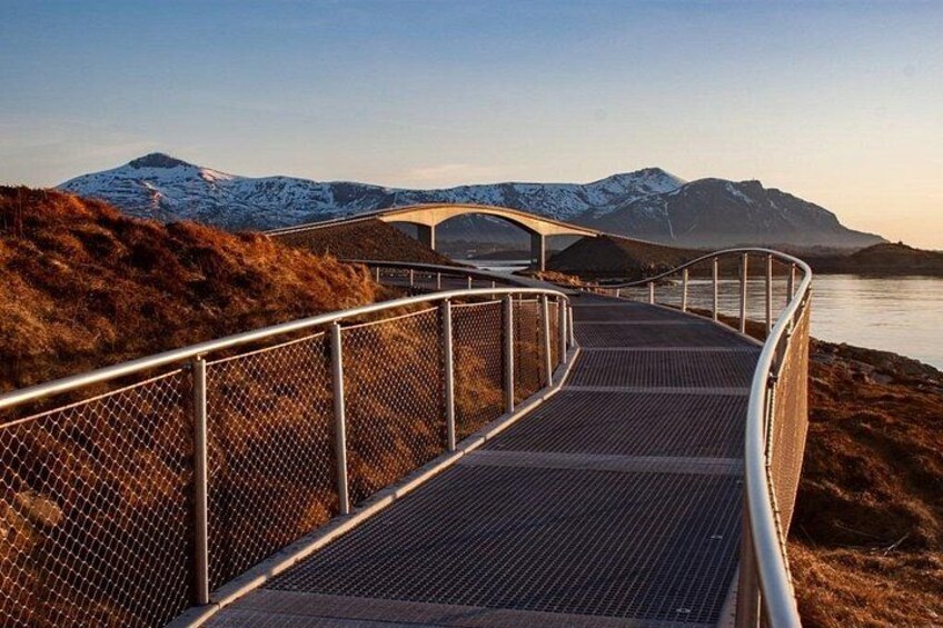 Eine Metallbrücke mit Metallgeländer, die zu einem Gewässer mit Bergen im Hintergrund führt. 2