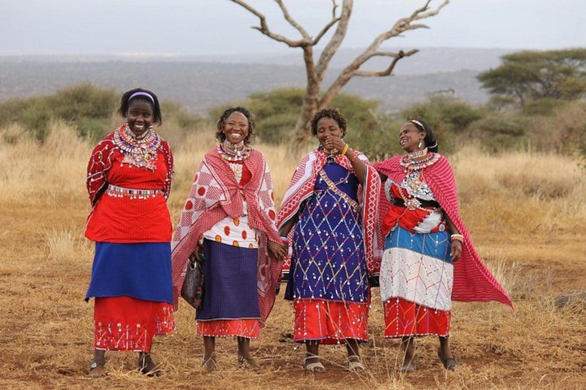 Four women in traditional attire standing in a grassy field with a tree and a distant landscape. 2