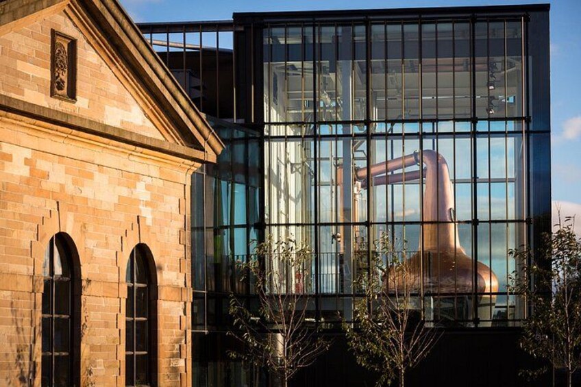 Ein Gebäude mit Glasfassade, das Industrieanlagen reflektiert, ein Backsteinbauwerk mit Rundbogenfenstern und ein klarer Himmel. 4