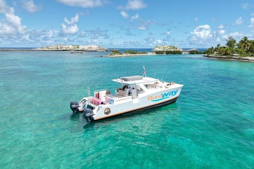 Een witte boot met de tekst 'Runaway' op de zijkant, drijvend in turquoise water onder heldere luchten en met land in de verte. 1