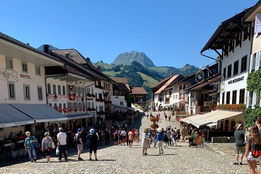 A cobblestone street in a European town with buildings labeled 'HOTEL' and 'RESTAURANT,' and mountains in the background. 5