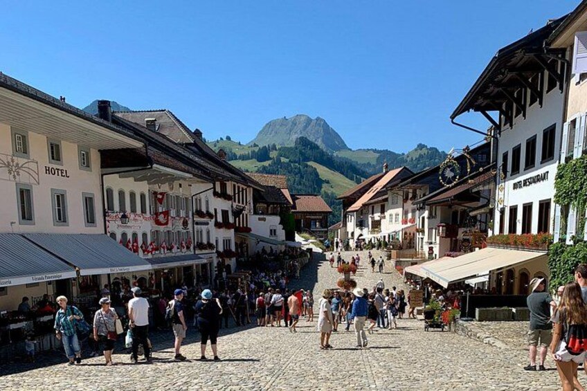 A cobblestone street lined with traditional buildings, a clock tower, and a mountain in the background. 2